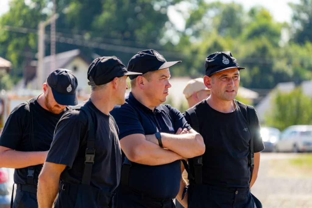 Firefighters in black uniforms and caps stand talking in a group on the square