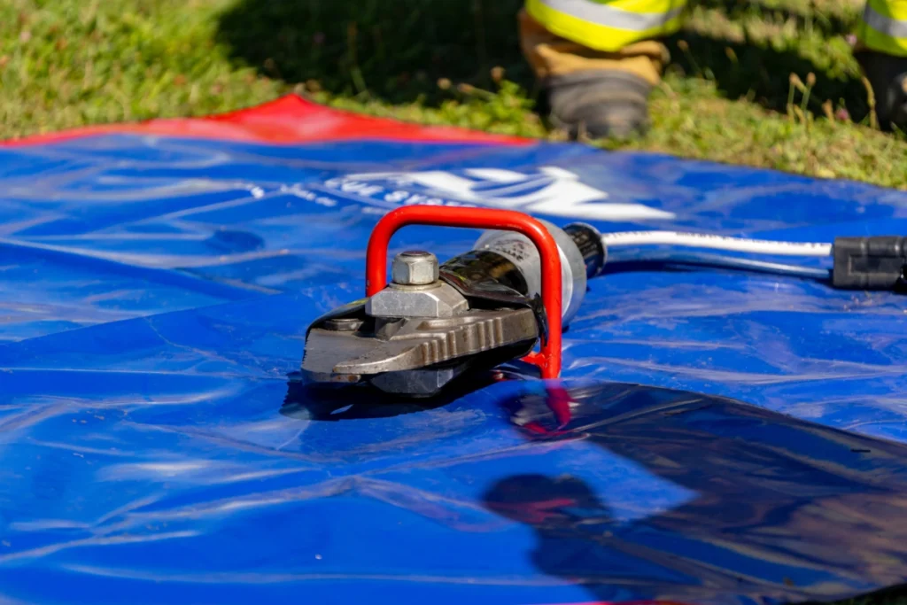 Hydraulic cutters with a red handle lie on a blue tarpaulin, a firefighter's boots visible beside them