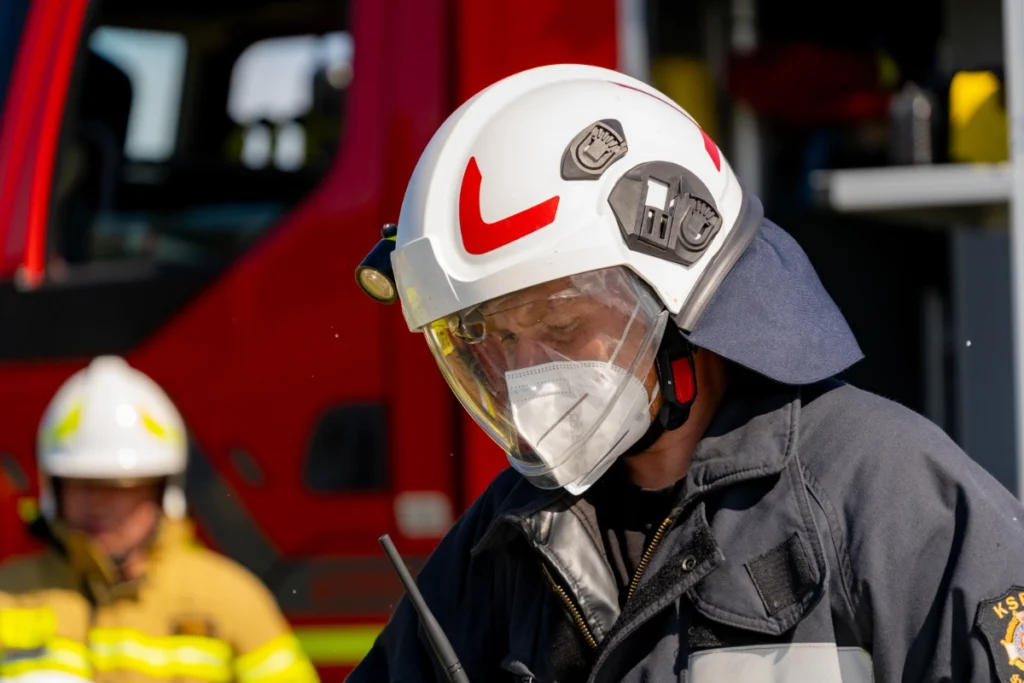 A volunteer firefighter in a white visored helmet and protective face mask bends down by a fire engine