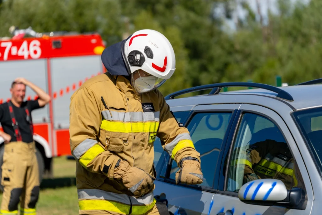 A volunteer firefighter in a sand-coloured uniform leans over a silver car's side window, fire engine behind