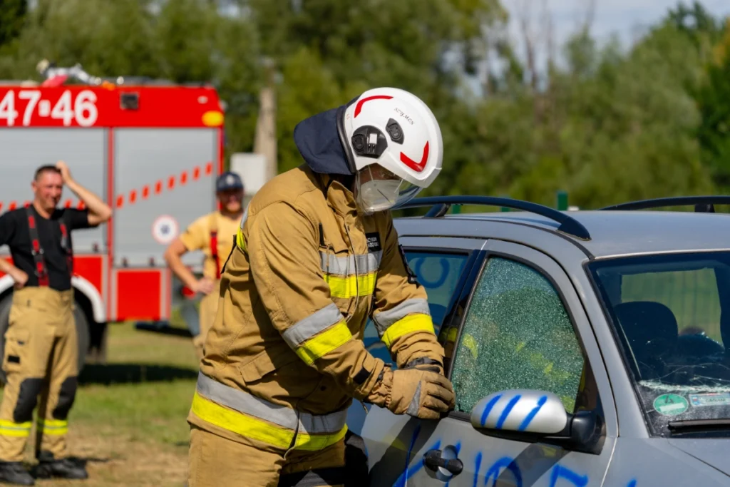 A volunteer firefighter smashes a silver car's side window, two firefighters standing by a fire engine behind