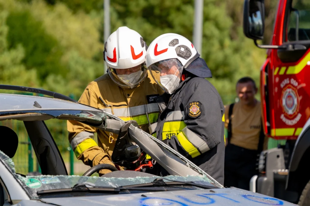 Two white-helmeted volunteer firefighters use hydraulic cutters on the pillar of a silver car with a smashed window