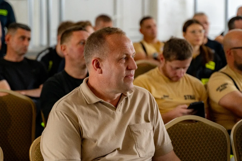 Participant in a beige polo shirt listens to the training, with uniformed volunteer firefighters seated behind him