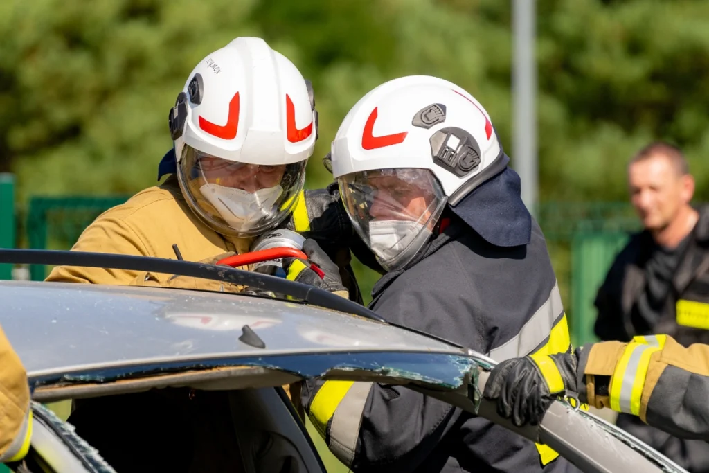 Two volunteer firefighters cut through a silver car's pillar with hydraulic cutters while a third steadies the roof