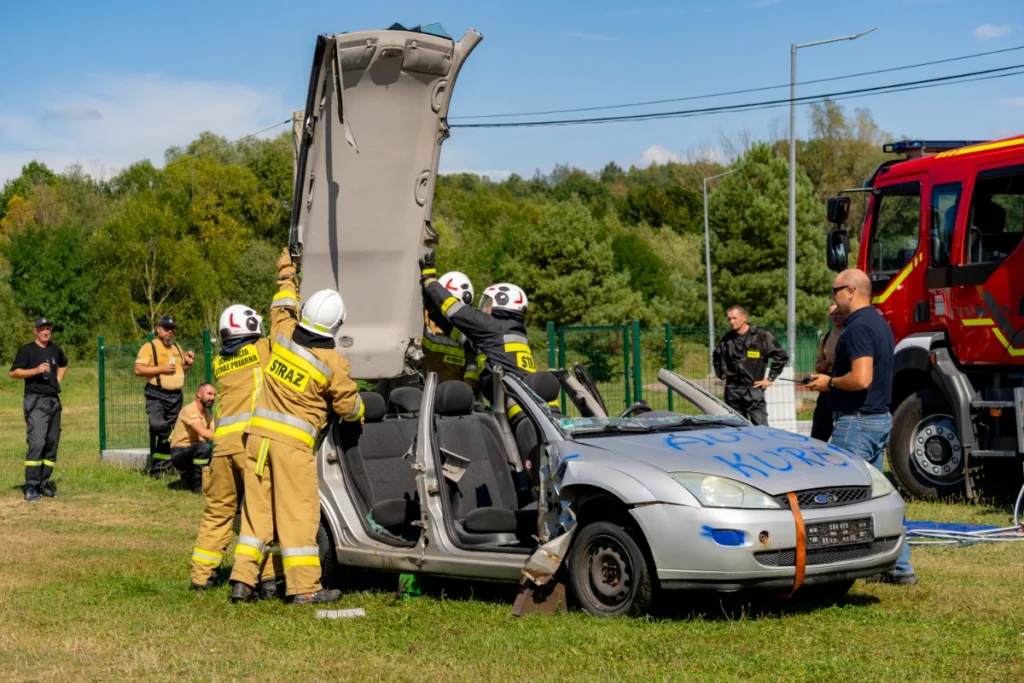 Volunteer firefighters raise upright the cut-off roof of a silver car marked AUTO KUREK, with spectators standing nearby