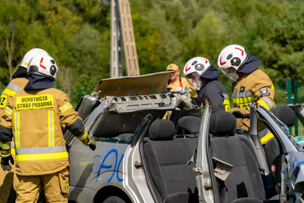 Volunteer firefighters peer into a silver car, its cut-off roof folded back onto the rear bodywork