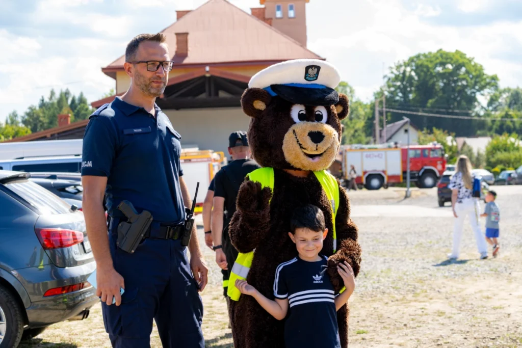A boy poses for a photo with a police bear mascot beside a policeman, fire engine behind