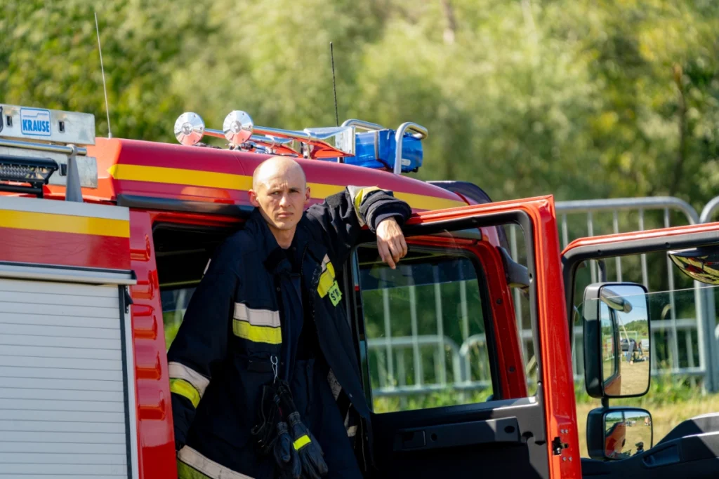 A volunteer firefighter in a dark uniform rests a hand on a fire engine's open cab door