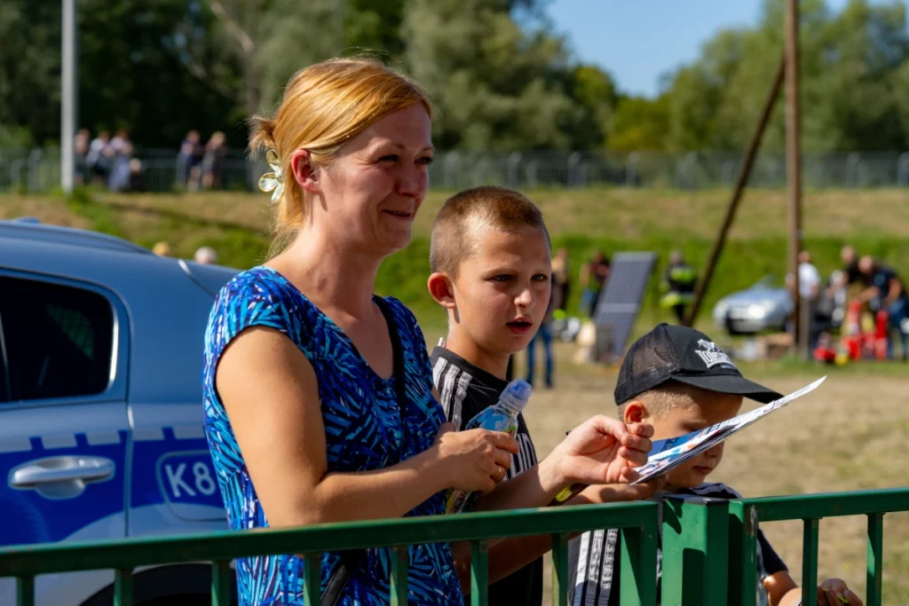 Woman and two boys watching from behind a green barrier, the younger holding a leaflet, police car nearby