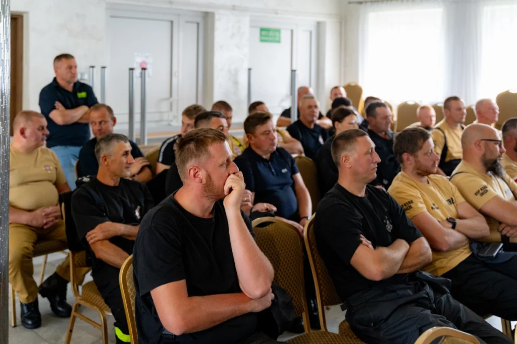Volunteer firefighters in sand-coloured and black uniforms fill the room, one standing at the back by the door