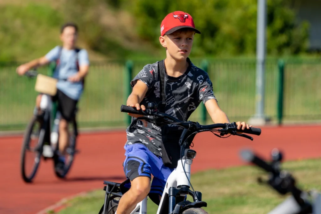 A red-capped boy rides a white electric bike, with a second cyclist on the red running track behind