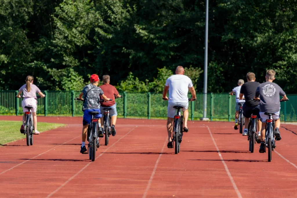 Seven people, children and adults, ride electric bikes along the red stadium running track, seen from behind