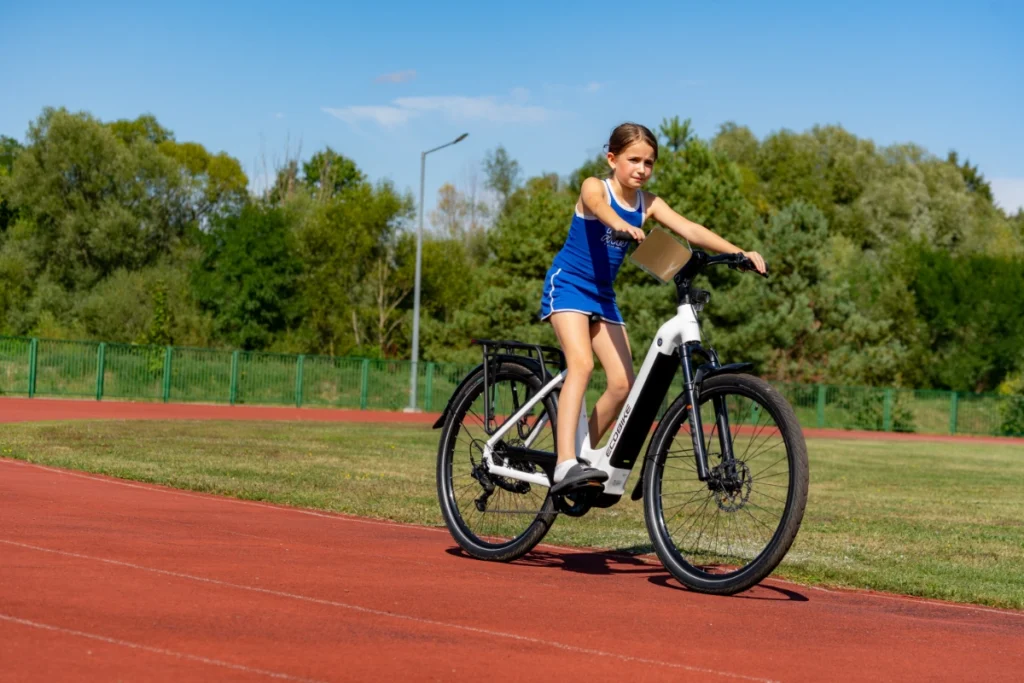 A girl in blue sportswear rides a white ECOBIKE electric bike along the red running track's edge