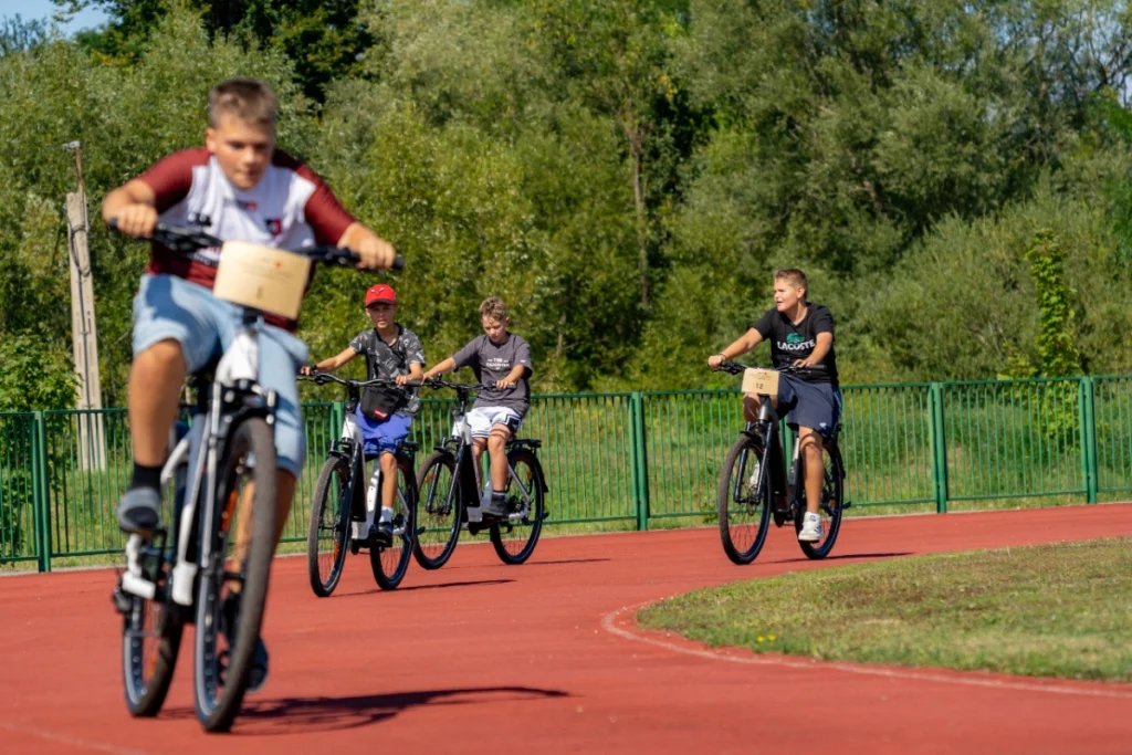 Four boys ride numbered electric bikes along the red running track, the first rider inches from the camera