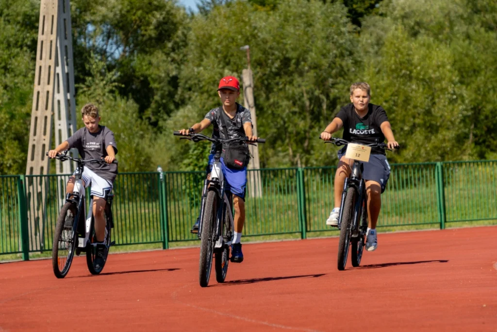 Three boys come riding electric bikes along the red stadium running track, one with number 12
