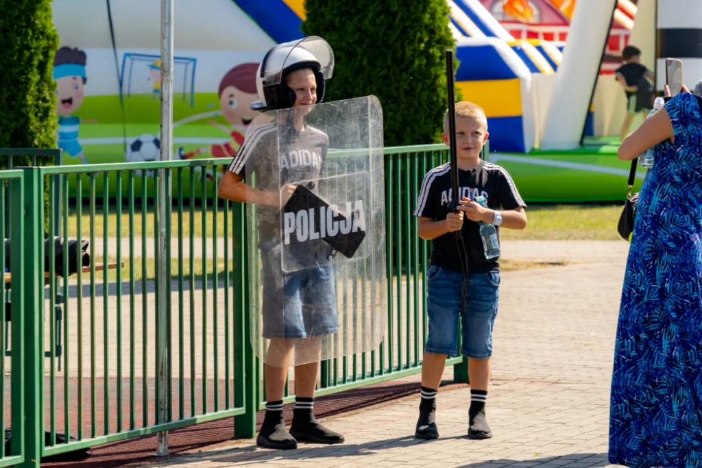 Two boys pose, one wearing a helmet and holding a shield marked POLICJA, the other a baton