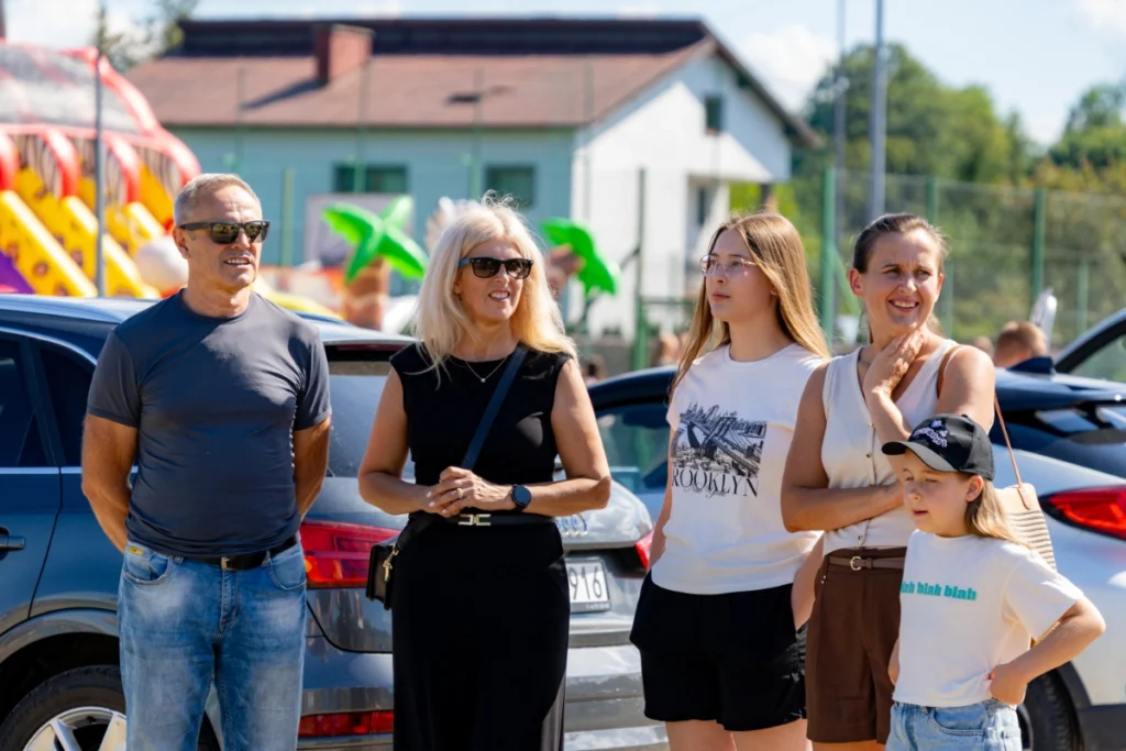 Picnic visitors, four adults and a girl, stand by parked cars with bouncy castles in the background