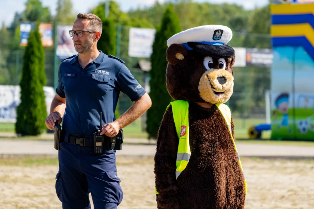 A uniformed policeman stands beside a police bear mascot in a white cap and reflective vest