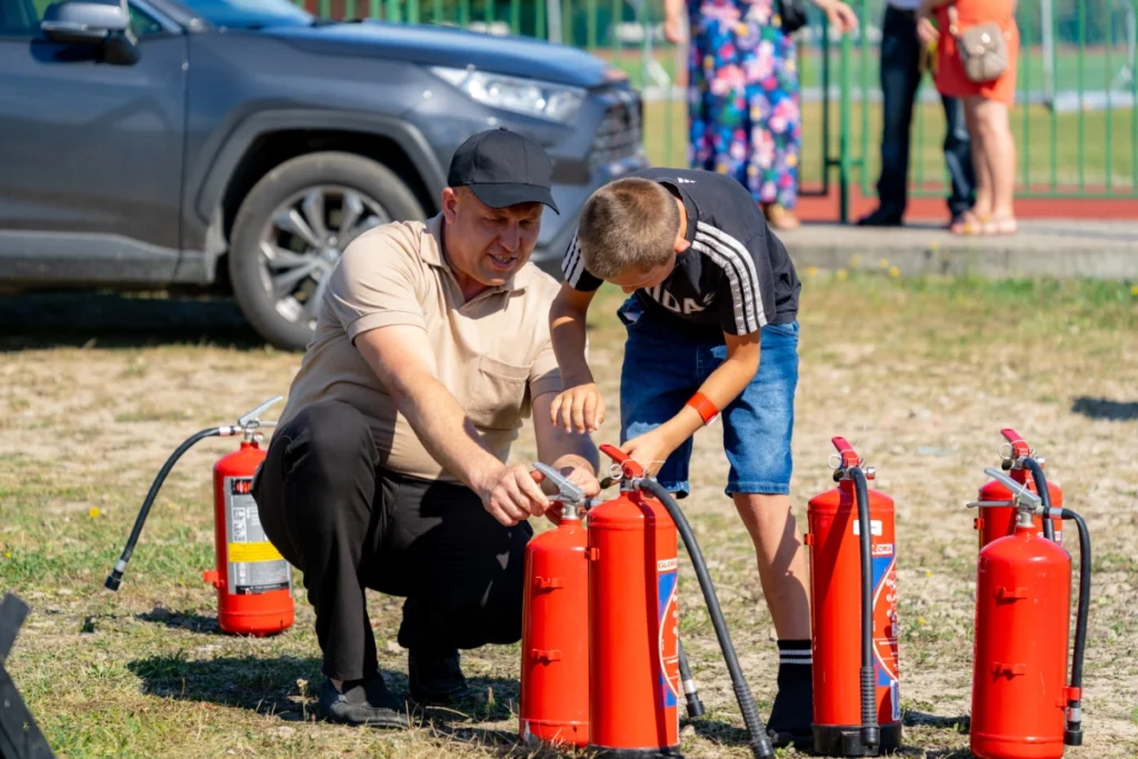 An instructor in a beige polo and a boy prepare one of several red extinguishers on the grass