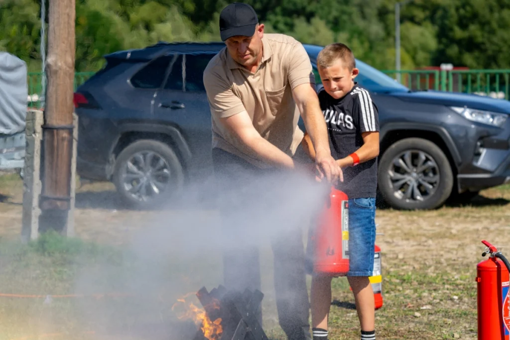 An instructor in a beige polo helps a boy douse a burning metal basket with a red extinguisher