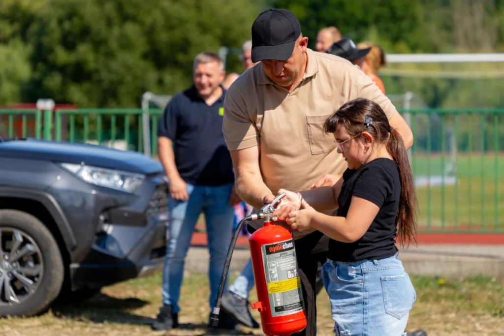 An instructor shows a girl how to hold a red fire extinguisher, picnic visitors watching in the background