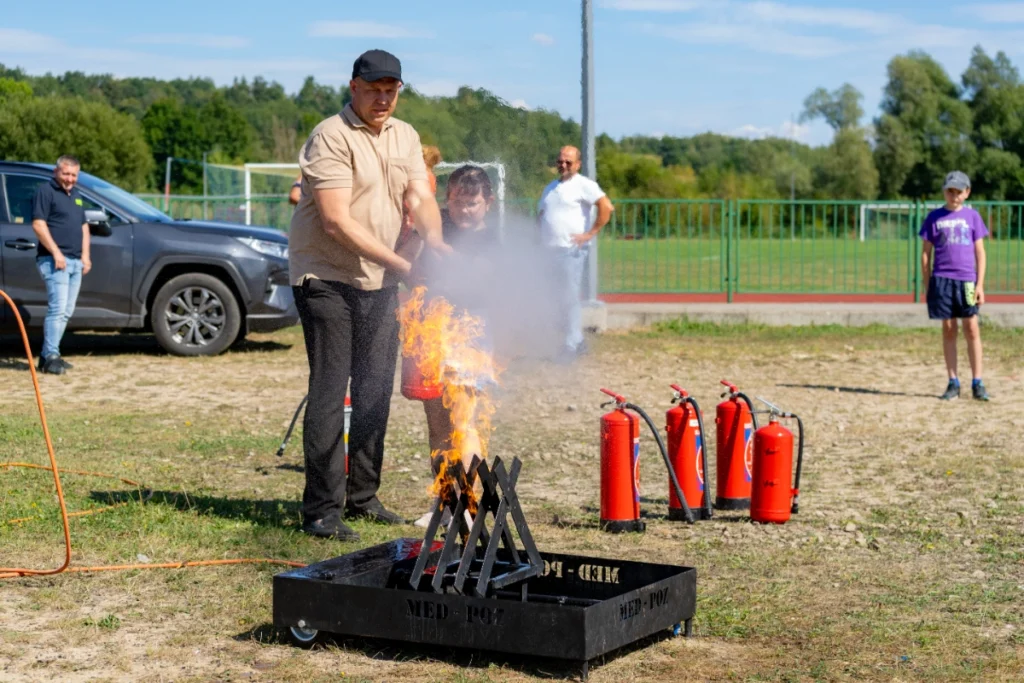 An instructor and a girl use a red fire extinguisher on flames in a metal basket marked MED-POŻ