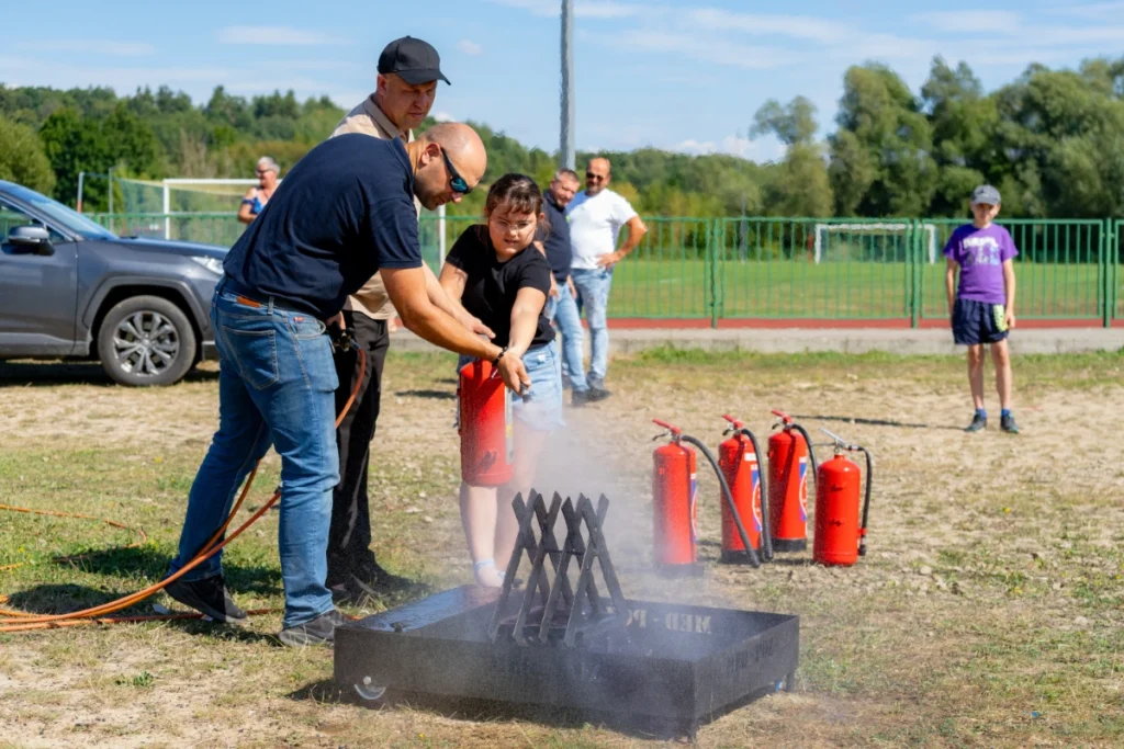 Two instructors help a girl douse a smoking metal basket with a red fire extinguisher, more extinguishers nearby