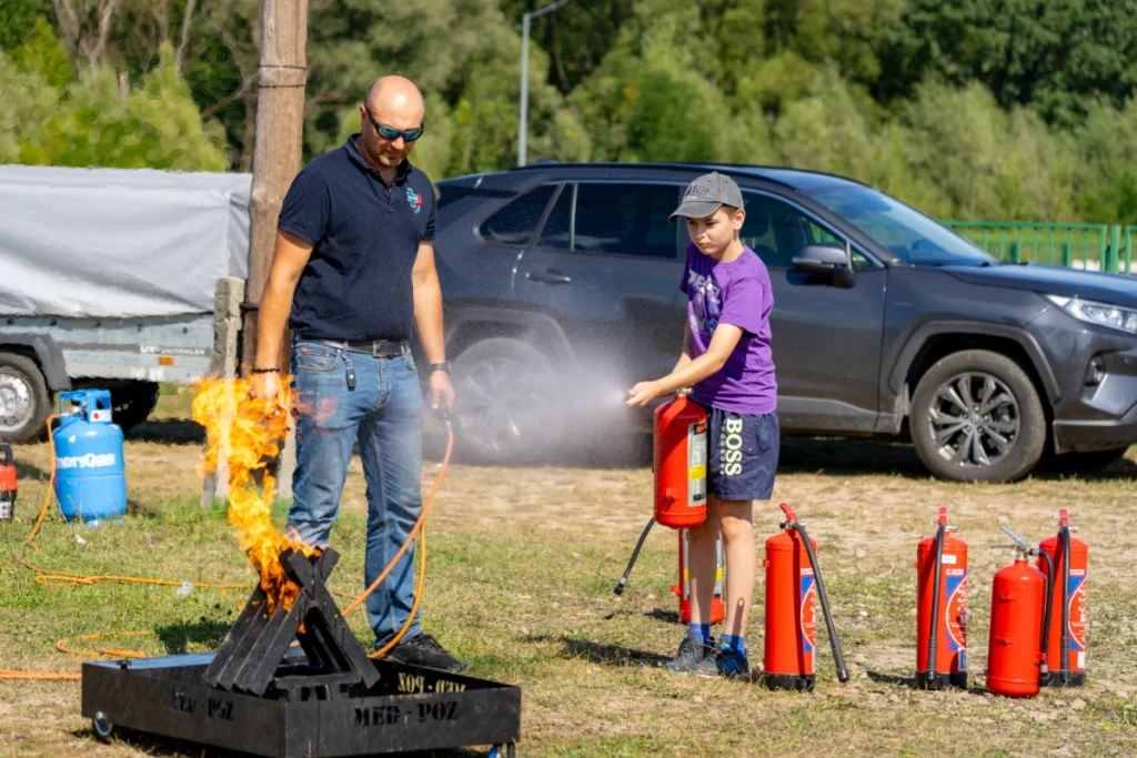 A purple-shirted boy douses a burning metal basket with a red extinguisher as a navy-shirted instructor stands by