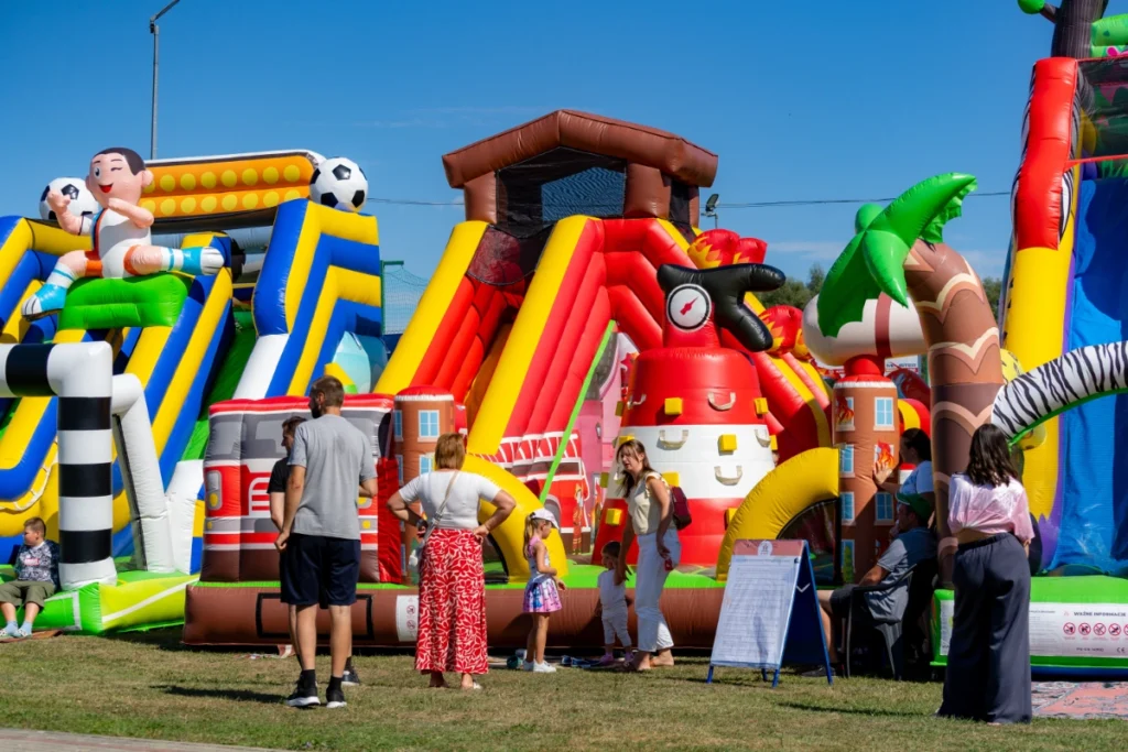 Families with children stand before large bouncy castles with slides, a footballer figure and a firefighting theme