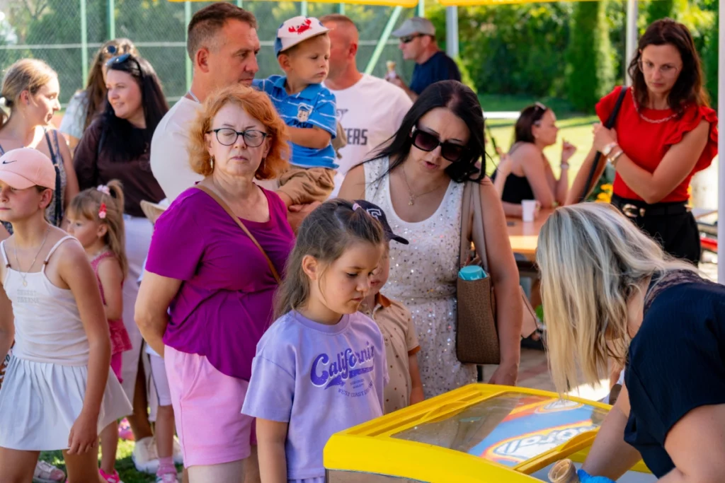 Children with their parents queue by a yellow freezer from which a saleswoman scoops ice cream