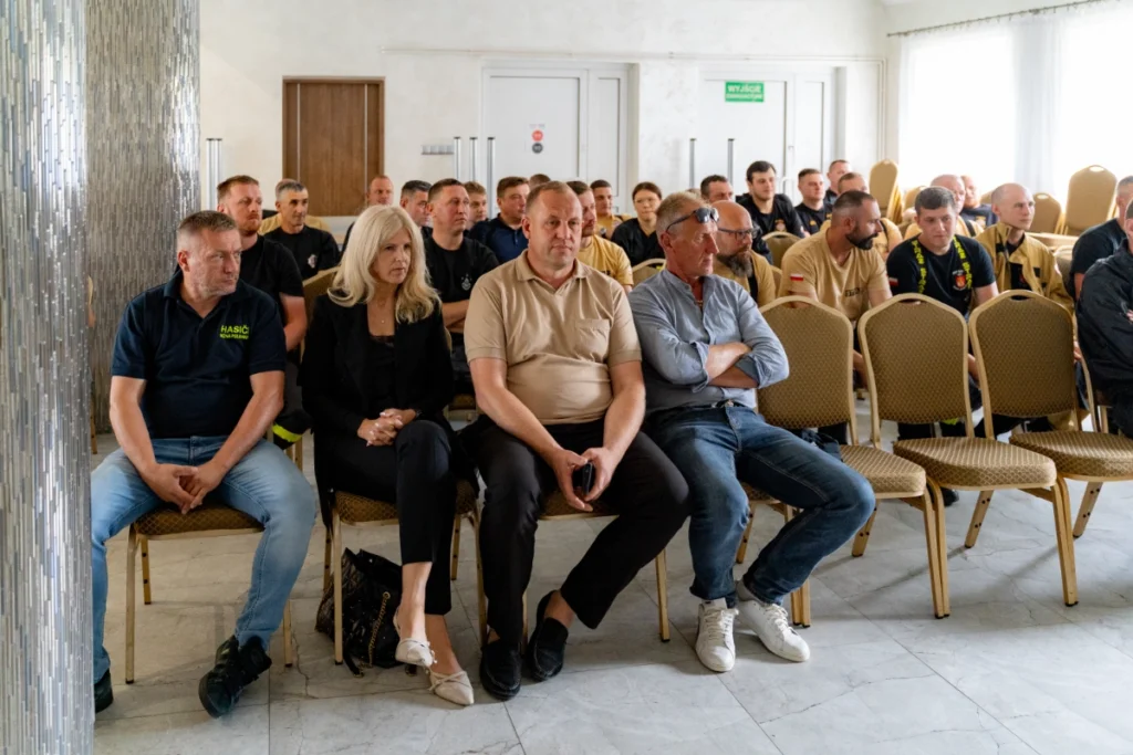 Firefighters and guests seated in rows of chairs, a man in a shirt reading HASIČI at the front