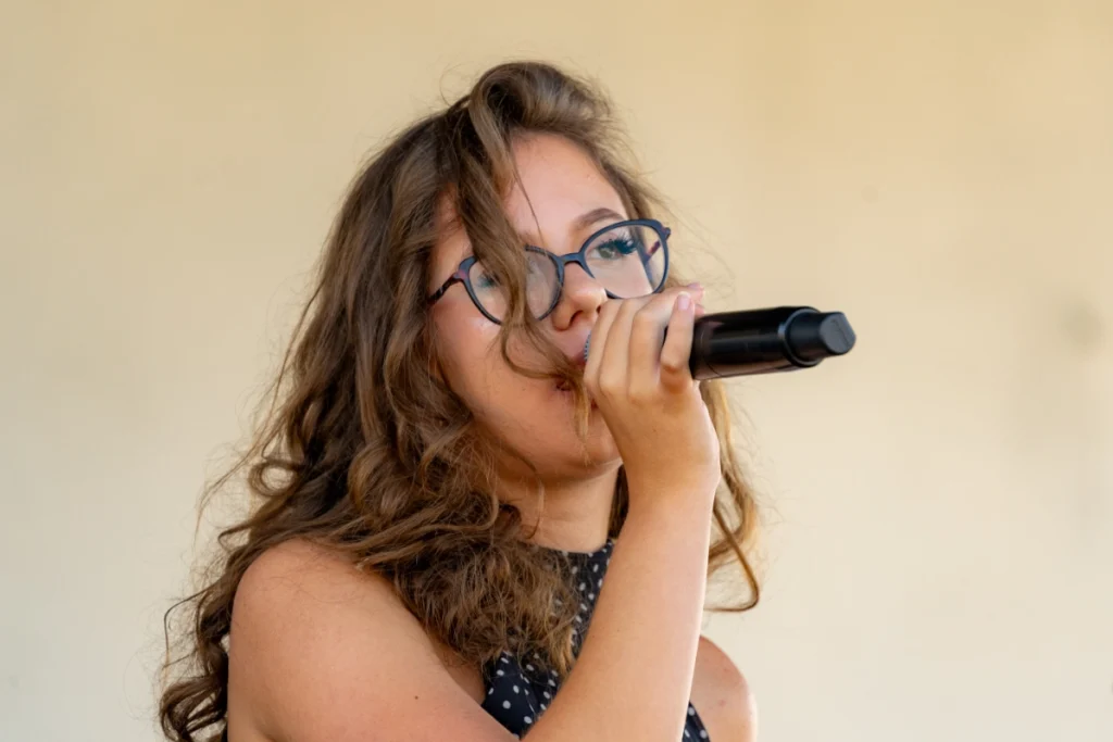 The youth band's vocalist sings with a microphone at her lips, in close-up against a pale stage wall