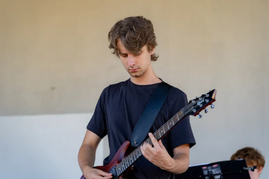On a covered stage, the youth band's guitarist plays a burgundy electric guitar, looking at the fretboard
