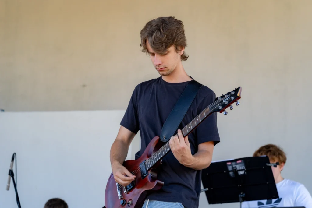 Guitarist playing a red electric guitar on a covered stage, keyboardist behind a music stand in the background