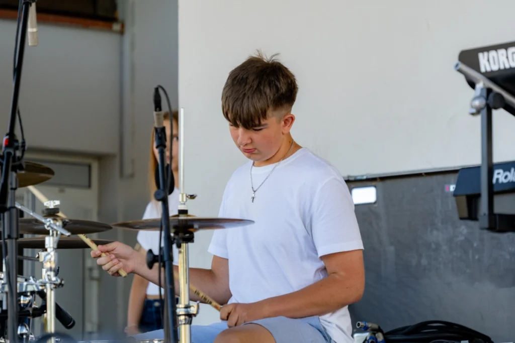 The band's drummer plays the drums with sticks on the covered stage, next to a keyboard
