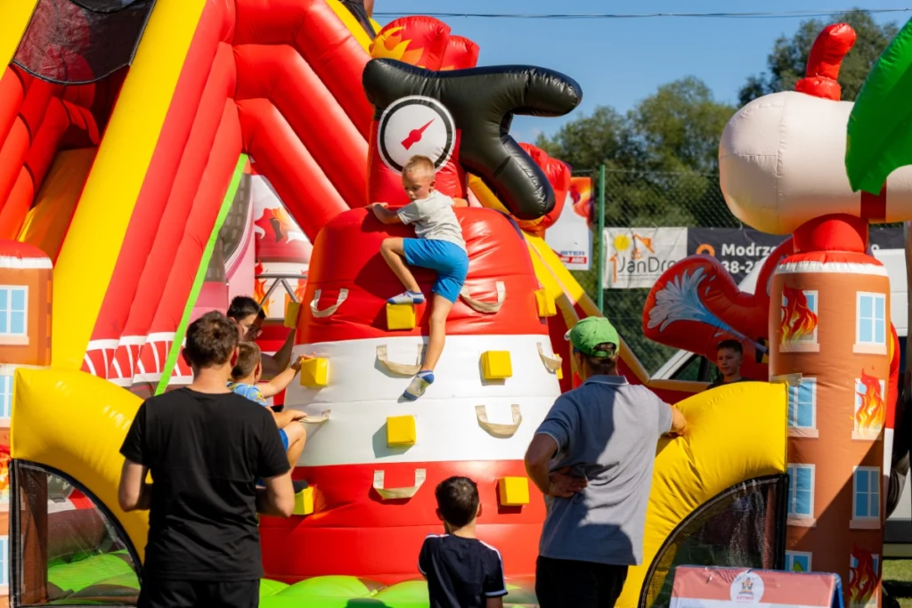 A boy scales the climbing wall on a firefighter-themed bouncy castle, children and adults standing nearby