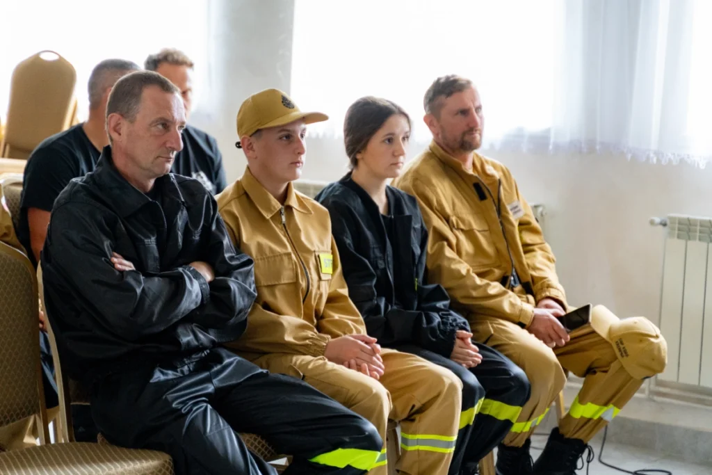 Volunteer firefighters in sand-coloured and black uniforms sit in a row and listen to the lecture