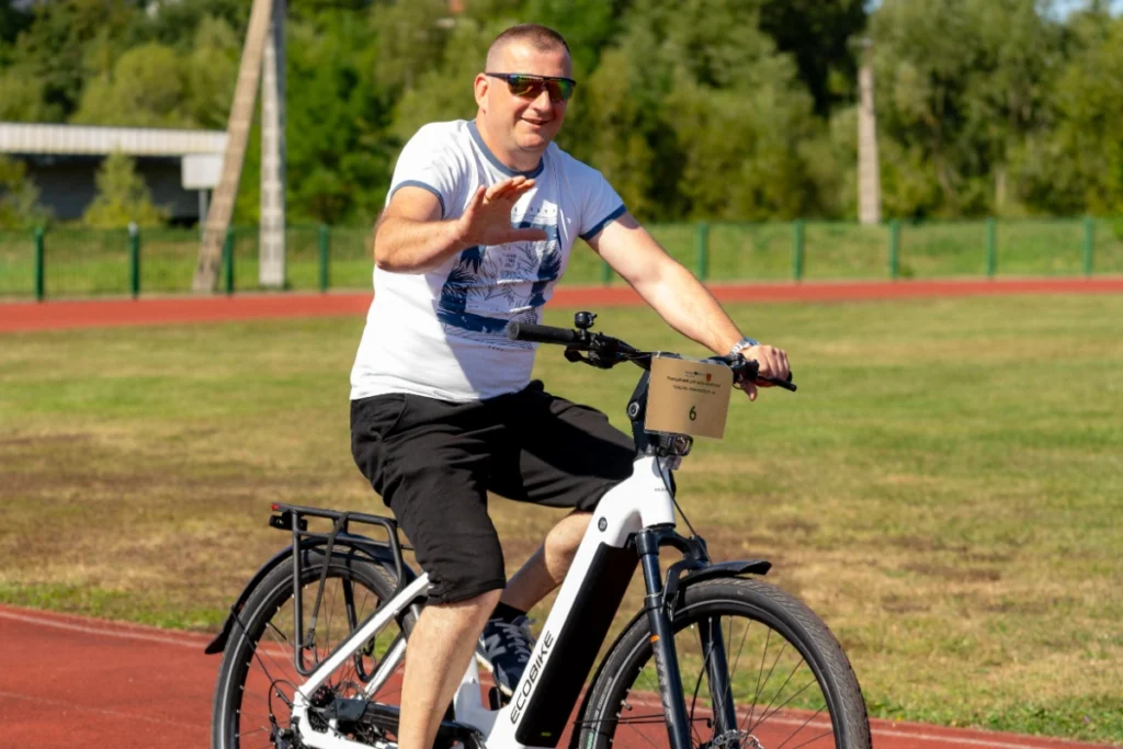 A man waves at the photographer while riding a white electric bike numbered 6 beside the red track