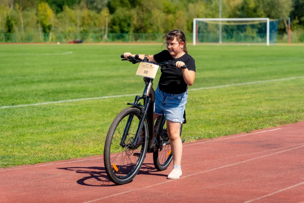 A girl wheels an electric bike numbered 10 along the red stadium running track, with the pitch behind