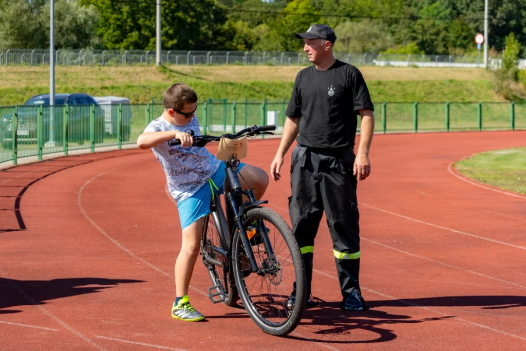 A boy sits on a graphite electric bike on the red stadium running track, beside a volunteer firefighter