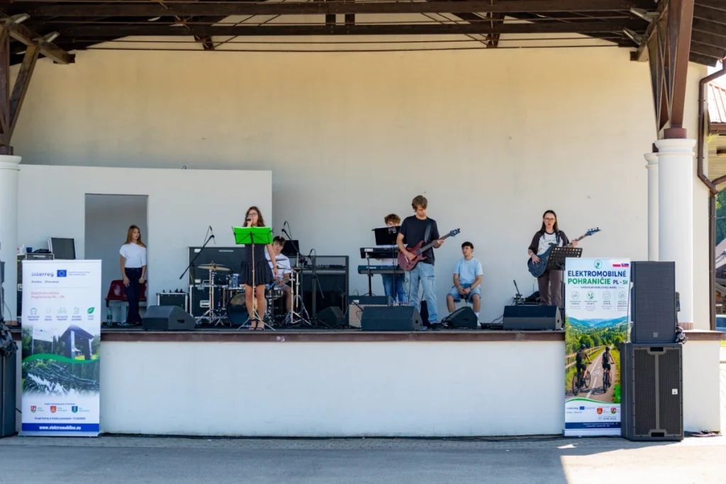 Youth band with a vocalist, guitars, drums and keyboard plays on a covered stage between project roll-up banners