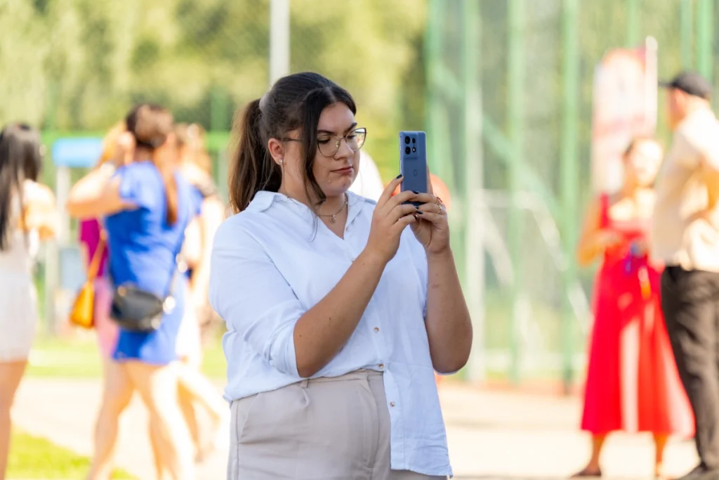 A picnic visitor takes a photo on her phone, with other visitors chatting by a green fence behind