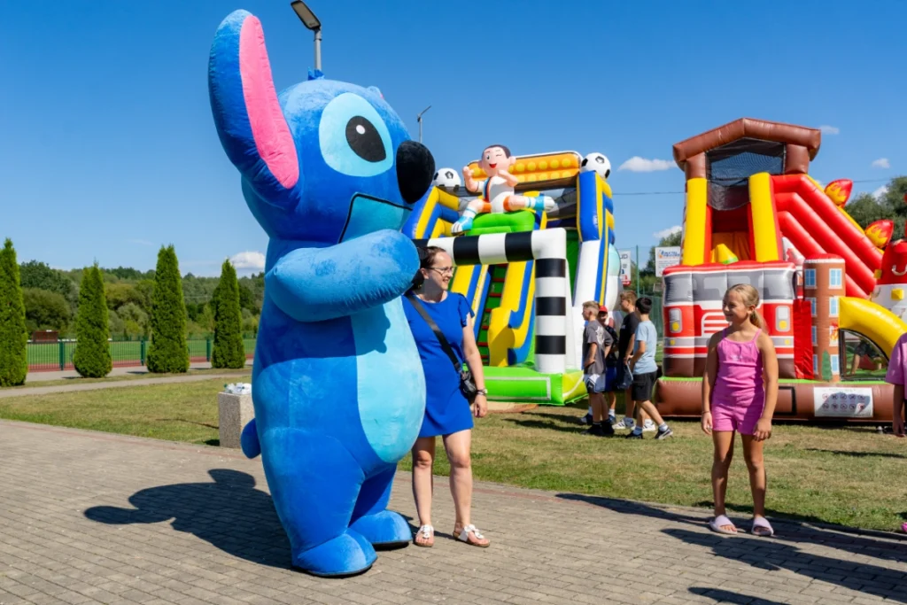 The Stitch mascot poses for a photo with a picnic visitor, a girl beside them, bouncy castles behind