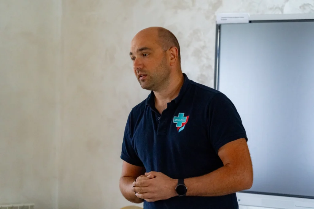 Training instructor in a navy polo shirt with the MED-POŻ logo speaks by a large screen