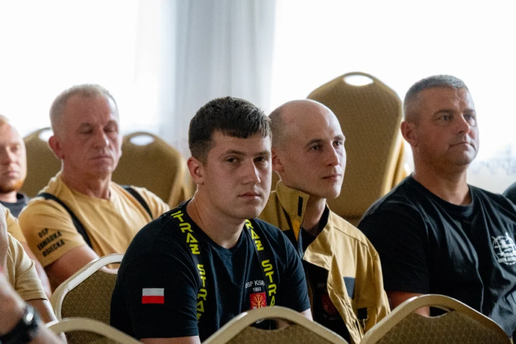 Volunteer firefighters in black and sand-coloured uniforms listen to the lecture, one shirt reading OSP ZAWADKA OSIECKA