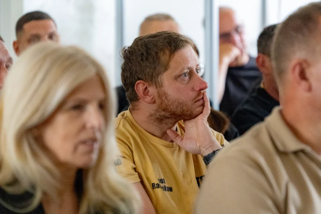 Firefighter in a sand-coloured shirt listens to the lecture, chin resting on his hand, beside other training participants