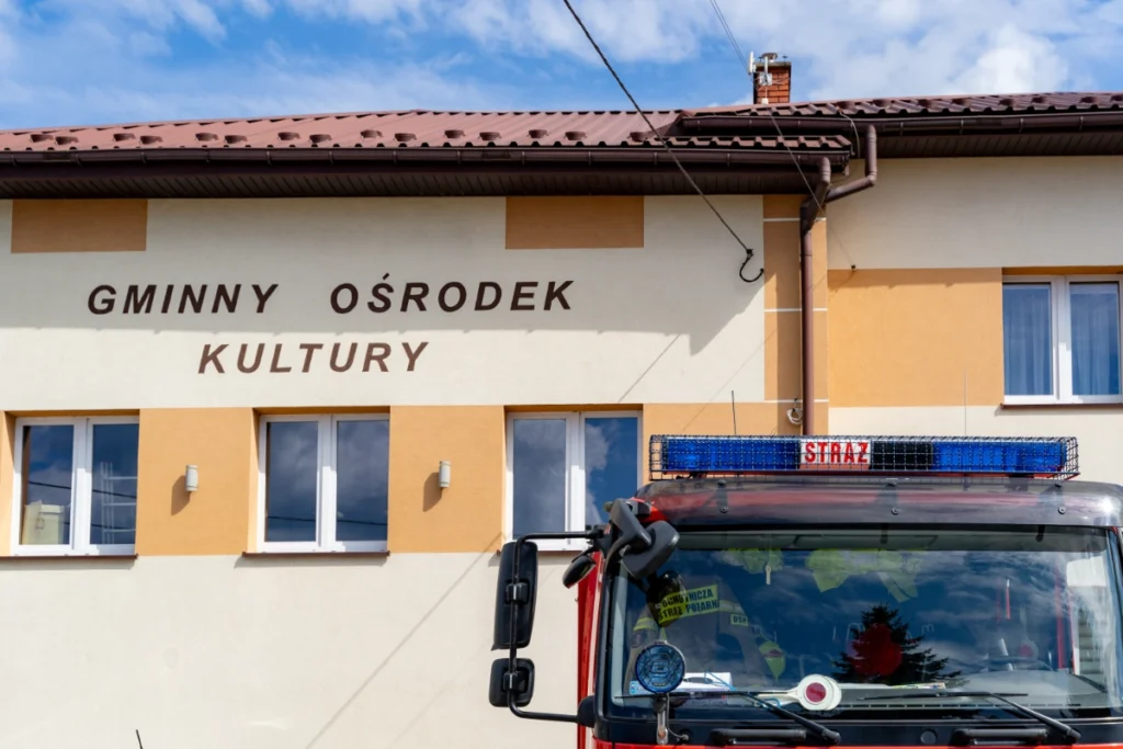 Facade sign reading GMINNY OŚRODEK KULTURY (Municipal Cultural Centre) behind a fire engine cab's STRAŻ light bar