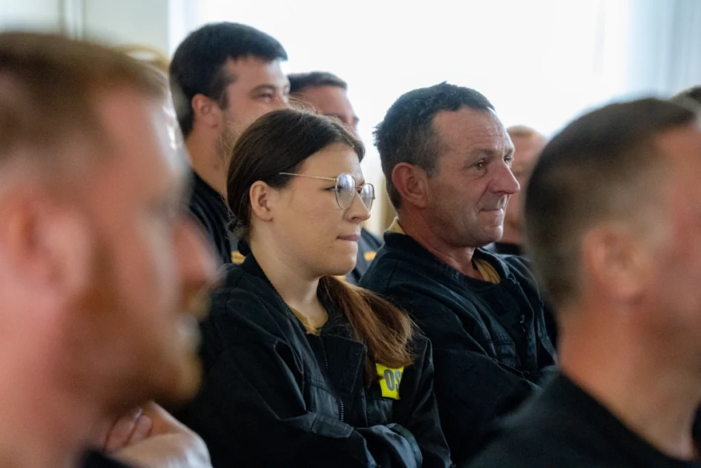A female and a male volunteer firefighter in black uniforms listen to the lecture, seated among other participants