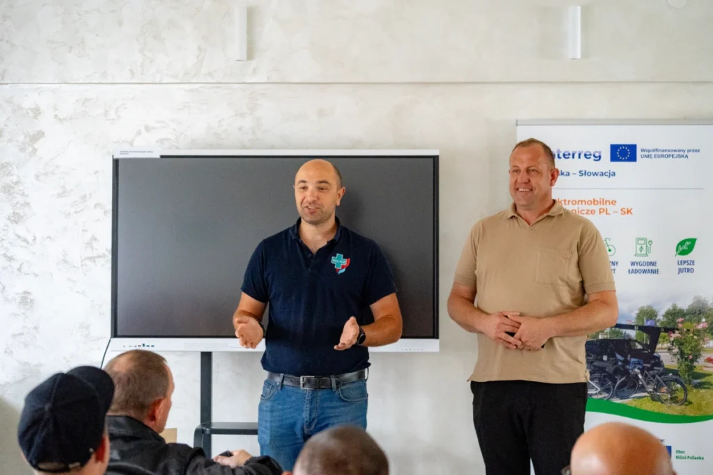 Training instructor speaks and gestures by the screen beside a man in beige at the Interreg roll-up banner