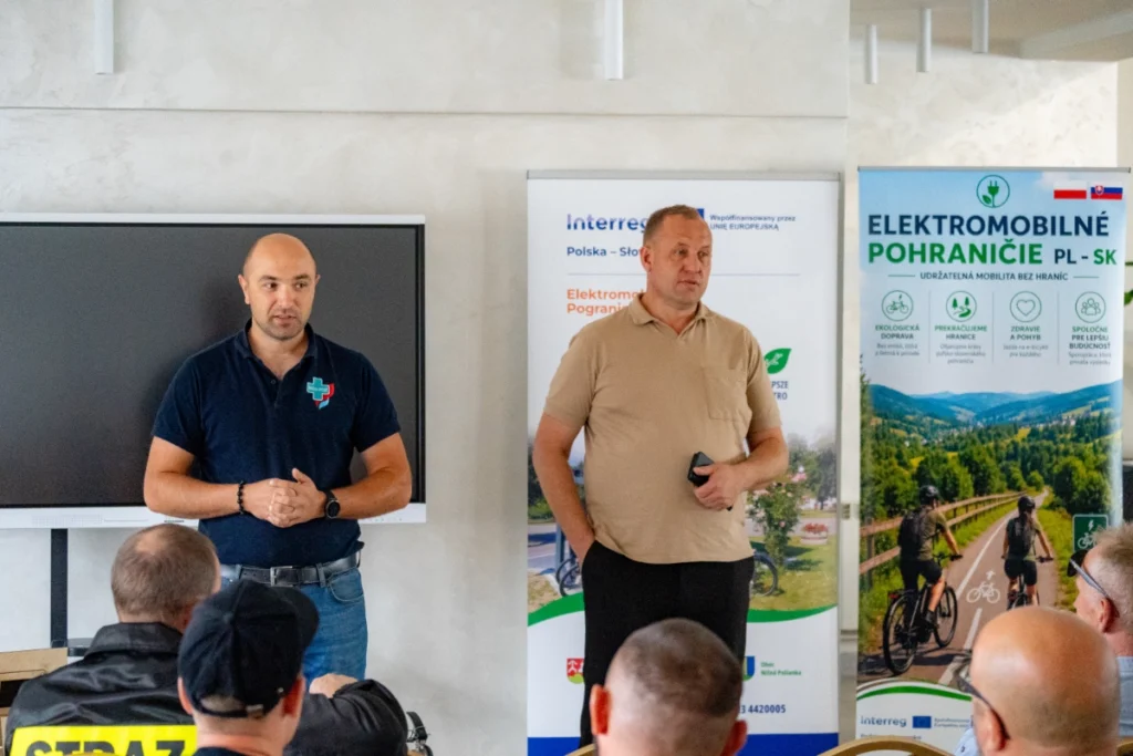 Man in a beige shirt addresses firefighters by the Interreg project roll-up banners, the training instructor beside him